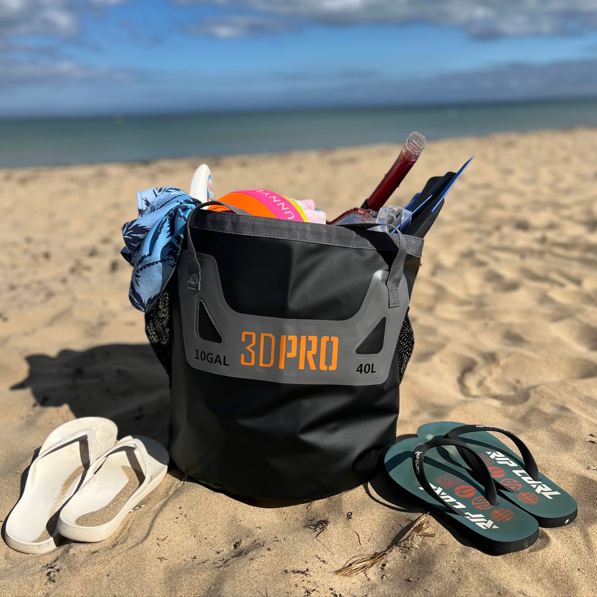 Black folding bucket with '3DPro' branding on a sandy beach with flip-flops and beach items.