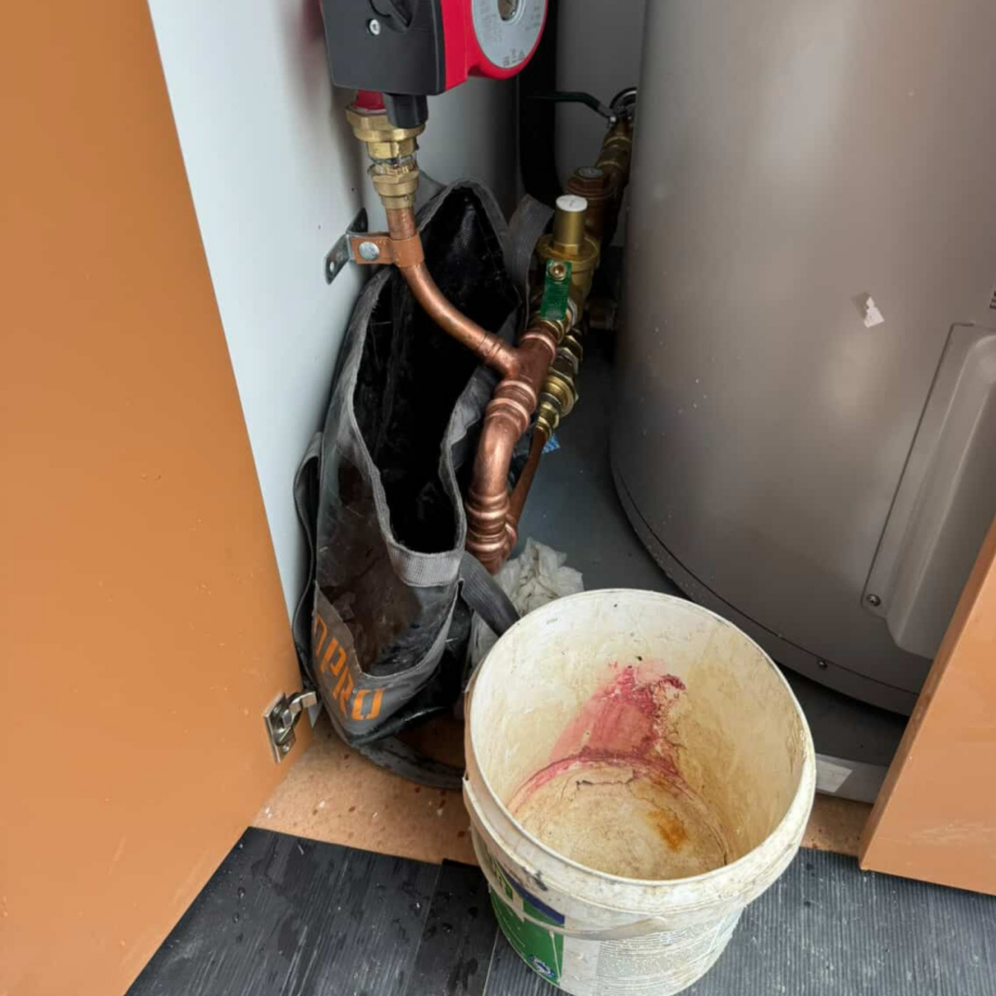 Plumbing work with pipes and a bucket on a wooden floor.