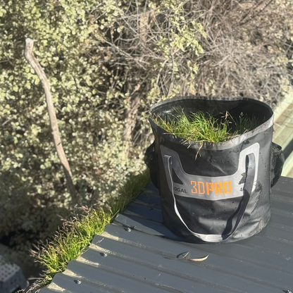 Black 3DPRO folding bucket bag with grass on a wooden surface, surrounded by greenery
