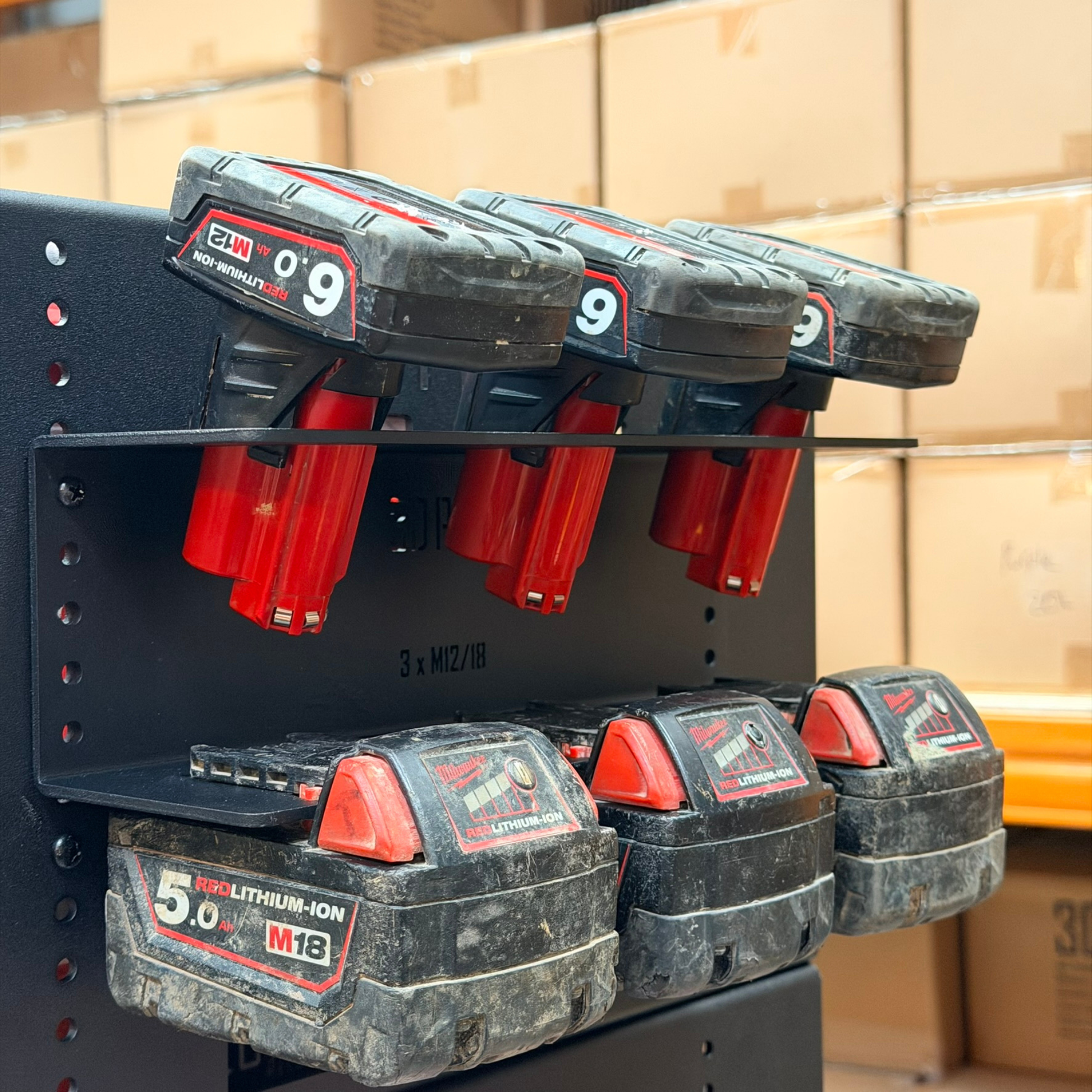 Set of Milwaukee M12 battery packs on a charging station with cardboard boxes in the background.